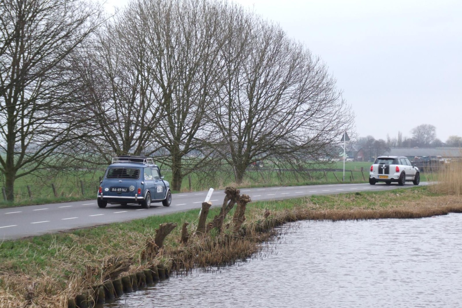 Een Mini Countryman R60 en een Mk1 rally Mini op een dijk langs een vaart met op de achtergrond kale bomen.
