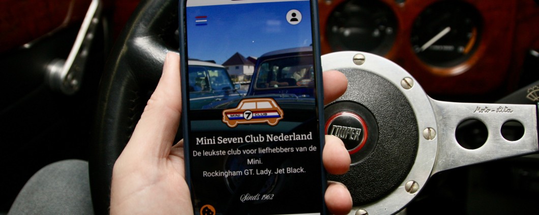 Hand met een mobiele telefoon waarop de nieuwe website te zien is met op de achtergrond het dashboard van een classic Mini